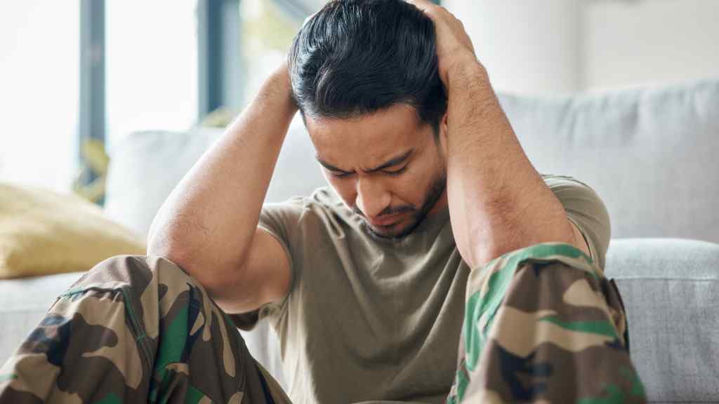 A man in a T-shirt and camouflage pants sits on the floor, holding his head in frustration. The living room setting suggests stress or contemplation.